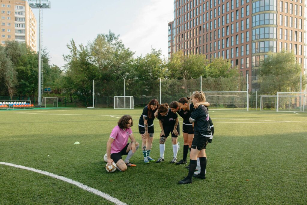 Women's soccer team in a huddle with coach on a sunny day, grass field.